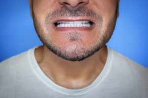 Parte baja de la cara de un hombre con barba que enseña los dientes de delante con la boca casi cerrada, sobre fondo azul