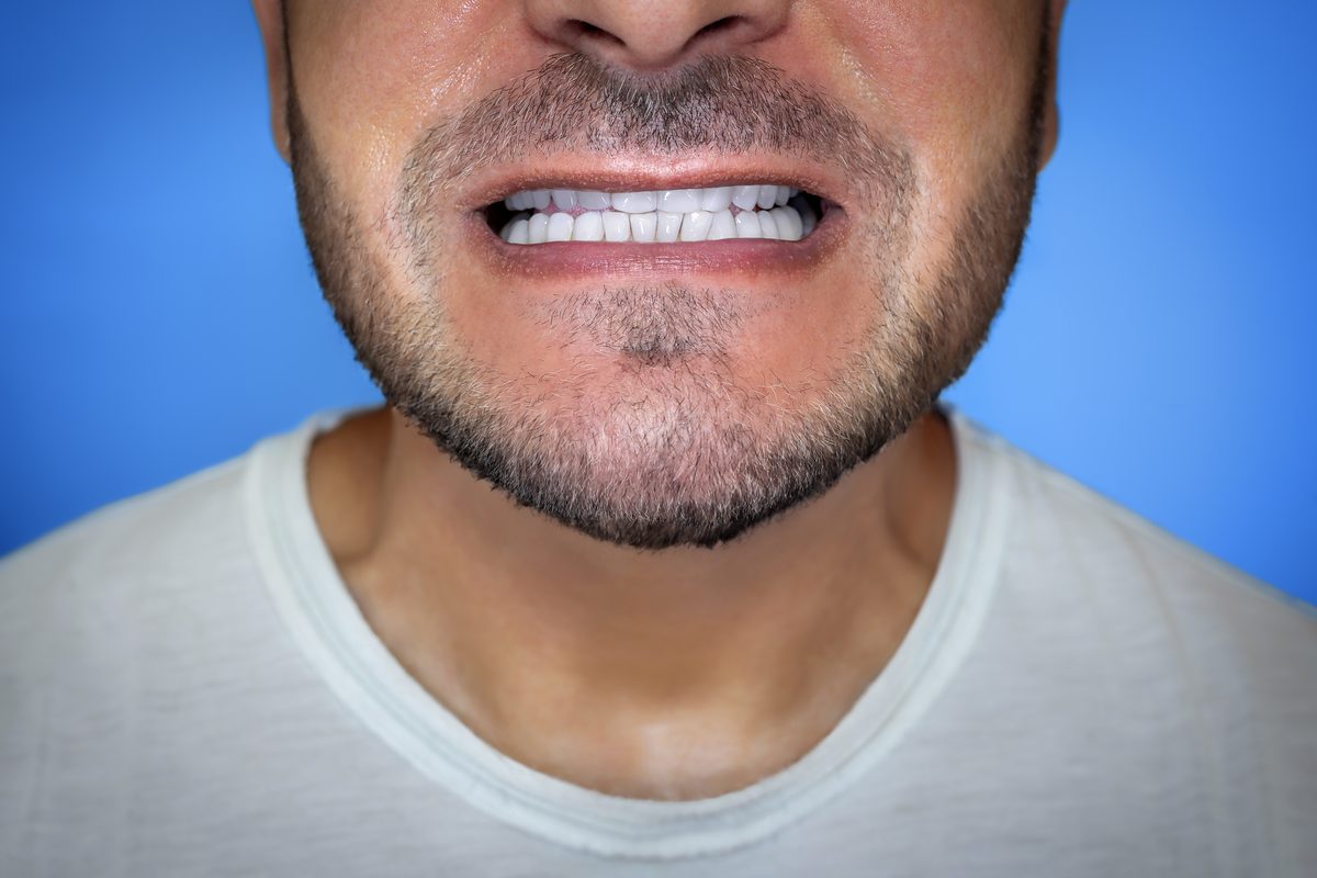 Parte baja de la cara de un hombre con barba que enseña los dientes de delante con la boca casi cerrada, sobre fondo azul